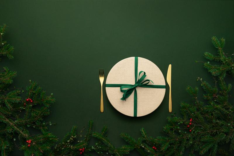 Vihreän taustan ympäröimänä on kuusenoksia. Kuvassa näkyy rusetilla ja lahjapaperilla paketoitu lautanen, jonka molemmilla puolilla kultaiset haarukka ja veitsi.