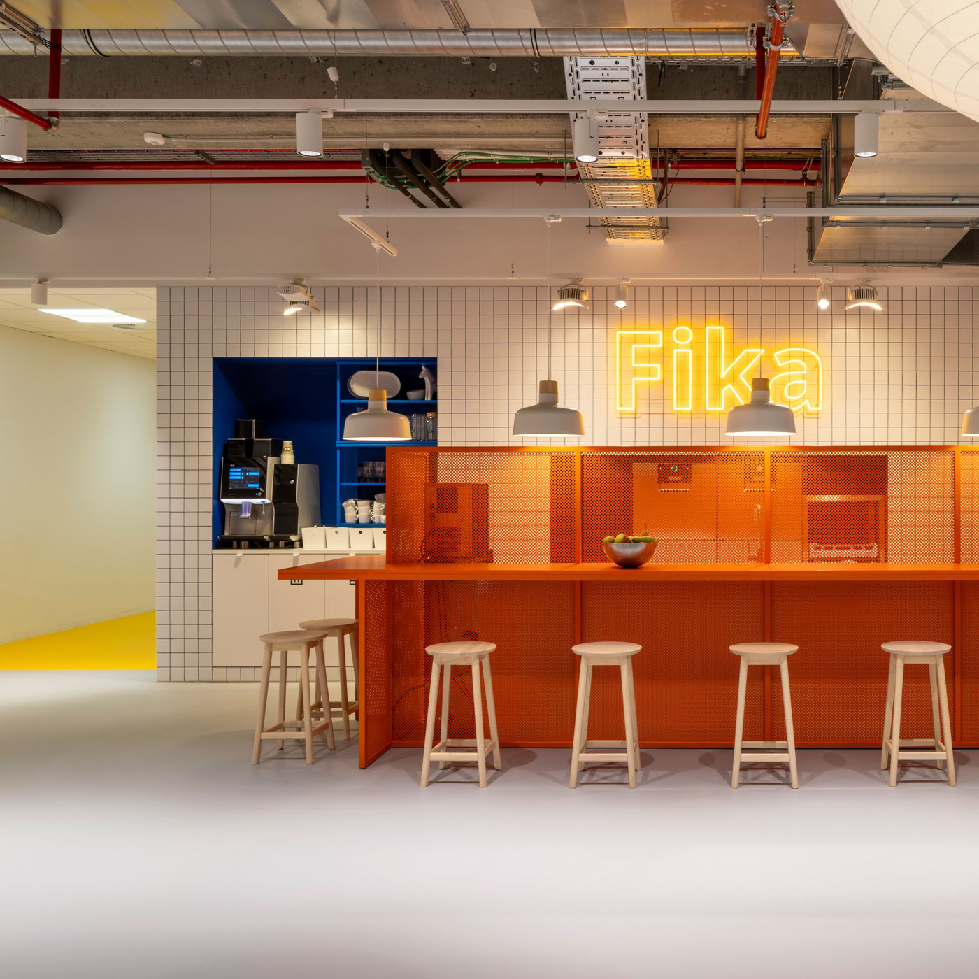 Un bar Fika orange vif avec des tabourets IKEA en bois clair et une enseigne néon lumineuse crée un coin accueillant pour les pauses café et les conversations informelles.
