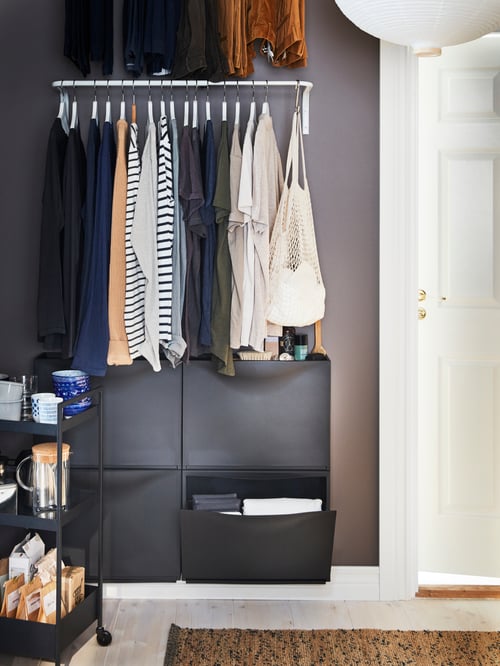 Clothing hangs on a bar mounted on a wall above four wall-mounted, black plastic TRONES shoe/storage cabinets by a door.