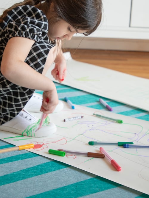 A girl sits on a large piece of paper, holding a MÅLA pen and drawing on her white sneakers.