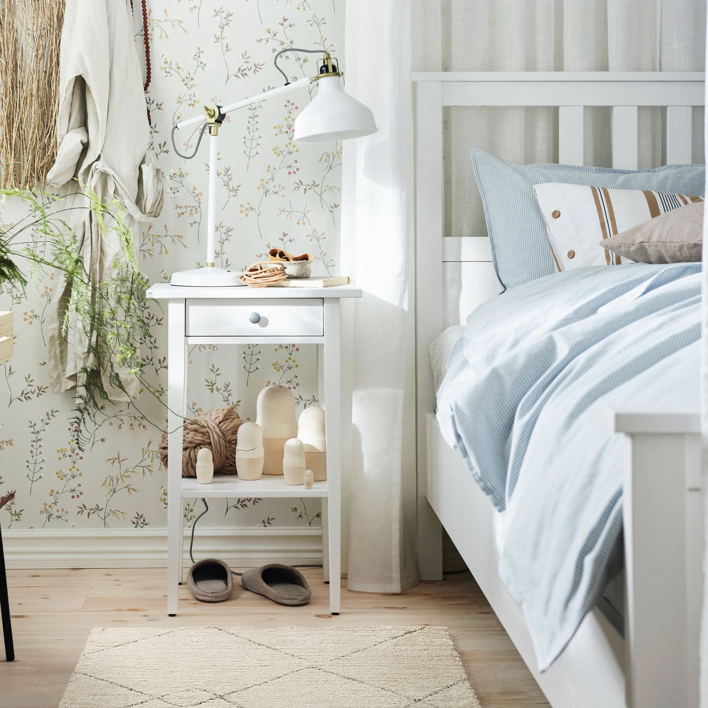 Ett ljust sovrum med en vit sängram och blårandigt bäddset. Bredvid sängen står ett vitt sängbord med låda och hylla, dekorerat med en vit RANARP skrivbordslampa, små träfigurer och växtdetaljer. Blommigt tapetmönster syns i bakgrunden.
