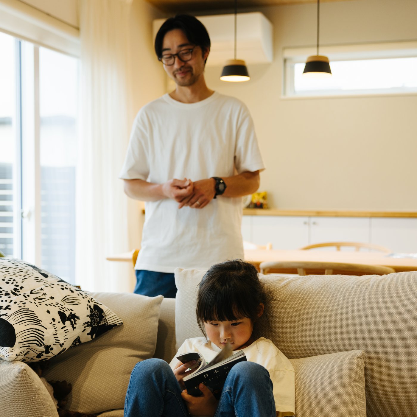 A father looks at his daughter who sits in a sofa reading a book.