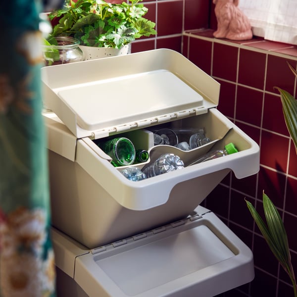A balcony featuring two beige SORTERA sorting bins with lid, filled with glassware and plastic bottles on a tiled balcony.