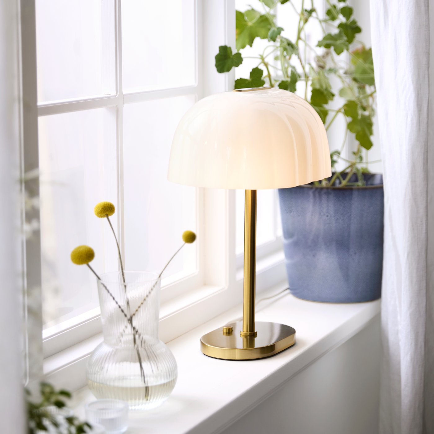 En opalvit AKTERSPRING bordslampa lyser upp ett mysigt inre bredvid en krukväxt och en dekorativ vas.