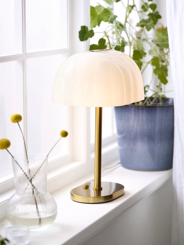 An opal white AKTERSPRING table lamp illuminates a cozy interior beside a potted plant and decorative vase.