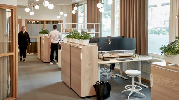 Bild på kontorslandskapet på Berzelii med flera arbetsstationer, stora fönster med bruna gardiner, två personer – en sittande vid dator och en gående. Datorskärmar står på skrivborden, krukväxter står utplacerade på olika ställen i rummet och hängande klotformade lampor hänger i taket.