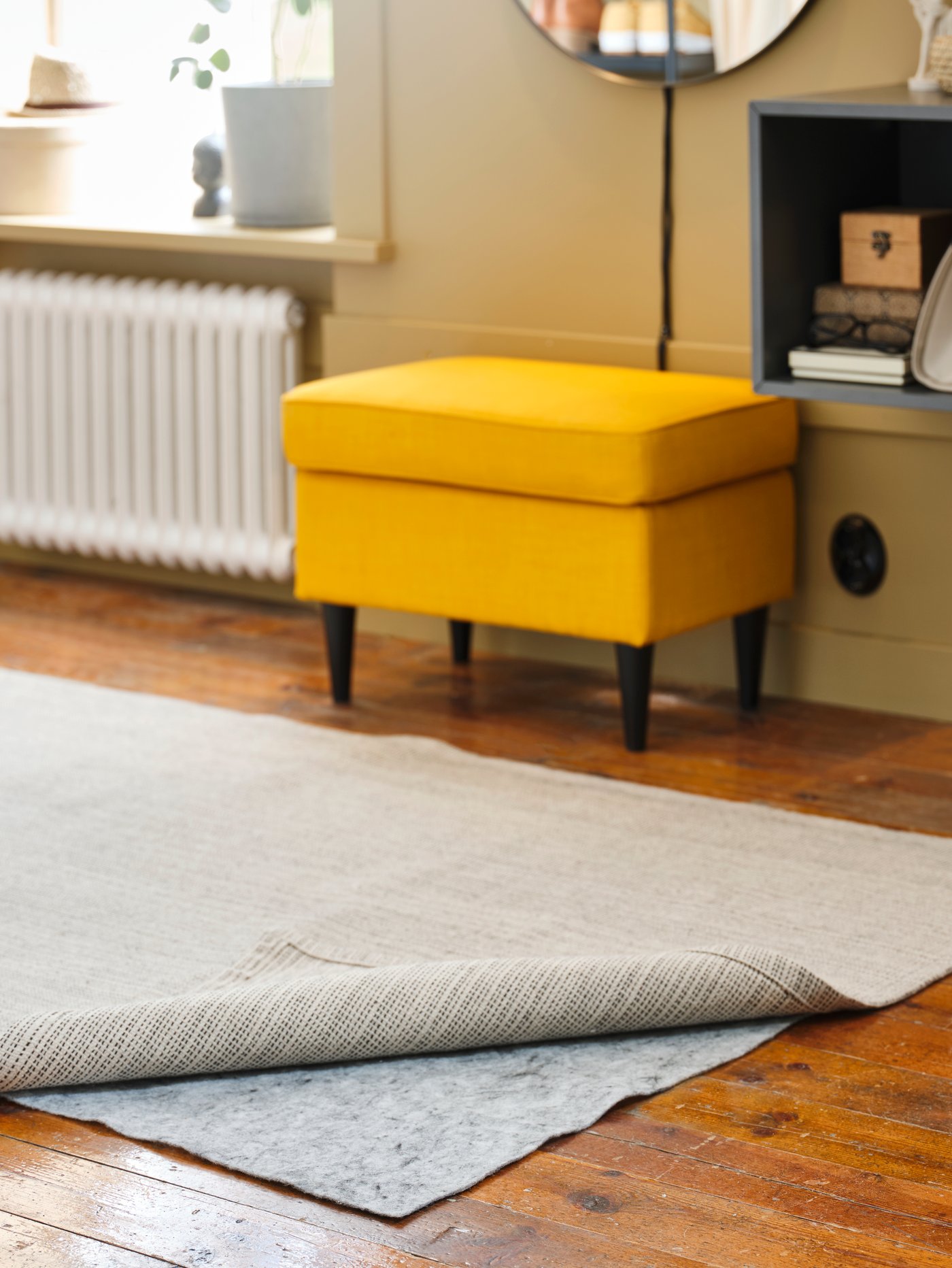 Bedroom area with a rug underlay with anti slip placed under a partially folded rug, with a yellow footstool to the side.