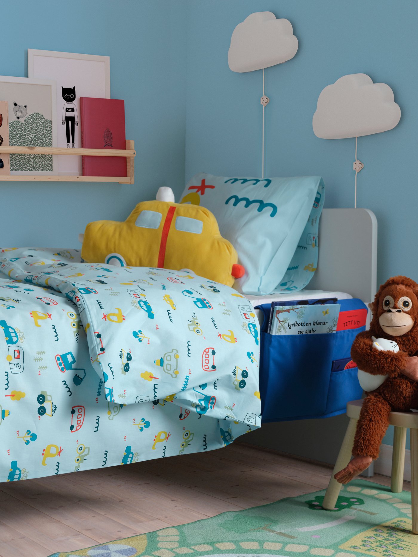 Een kleurrijke IKEA kinderkamer toont een bed versierd met lichtblauw beddengoed met voertuigpatronen en een geel autovormig kussen, aangevuld met een houten BEKVÄM kruidenrek dat boeken bevat, twee wolk-vormige UPPLYST wandlampen, een blauwe bedorganizer en een DJUNGELSKOG knuffelaap op een klein krukje.