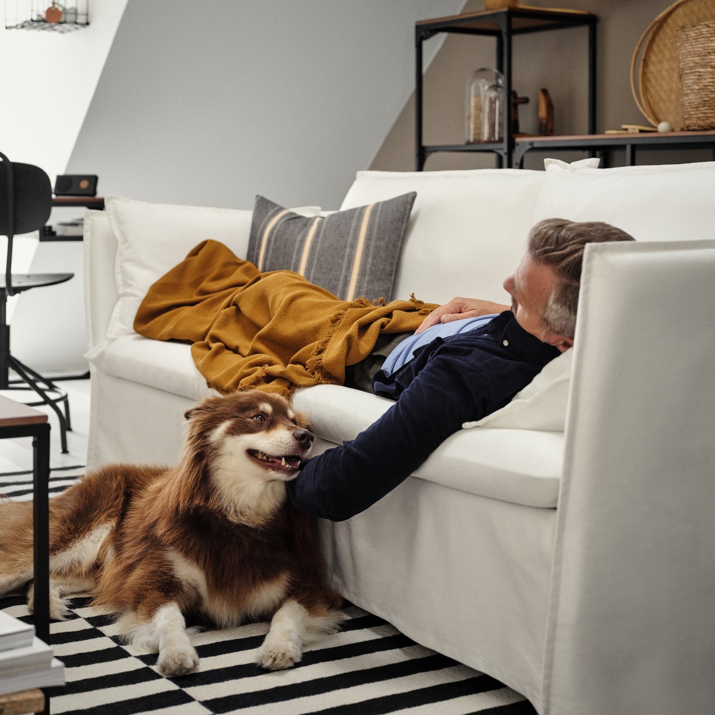 A person is laying on a white couch in a living room petting a dog who is laying on a striped rug.