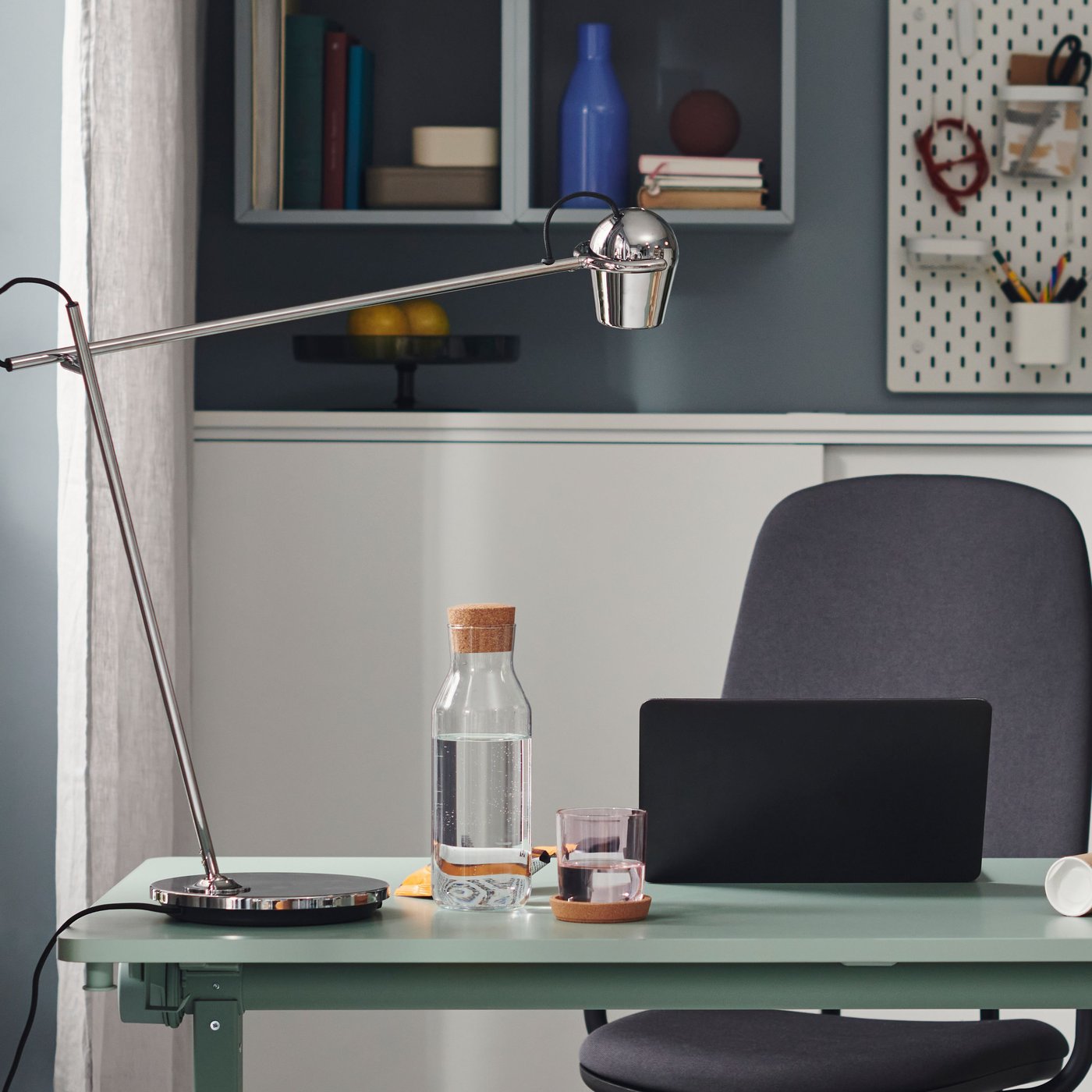 그린 색상의 접이식 테이블 위에 크롬 도금 MODERMOLN 모데르몰른 독서등이 놓여있고 조명의 물방울 모양 헤드가 노트북 위로 기울어져 있는 모습.