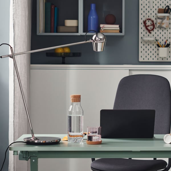 A chrome-plated MODERMOLN work lamp stands on a green foldable table. The lamp’s drop-shaped head is angled over a laptop.