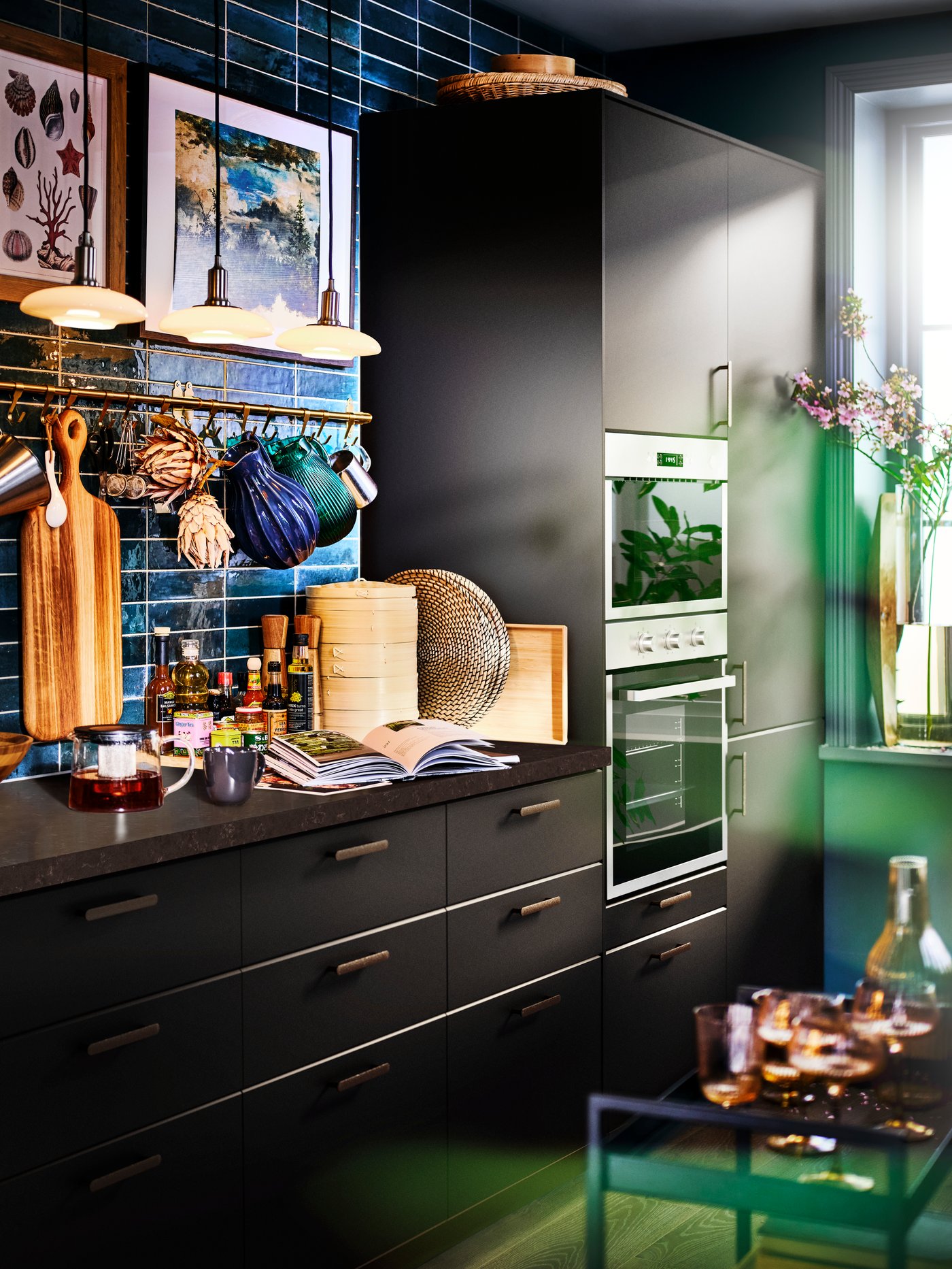 A KUNGSBACKA anthracite kitchen with a blue-green tiled wall, with pendant lamps above a worktop with kitchen accessories.