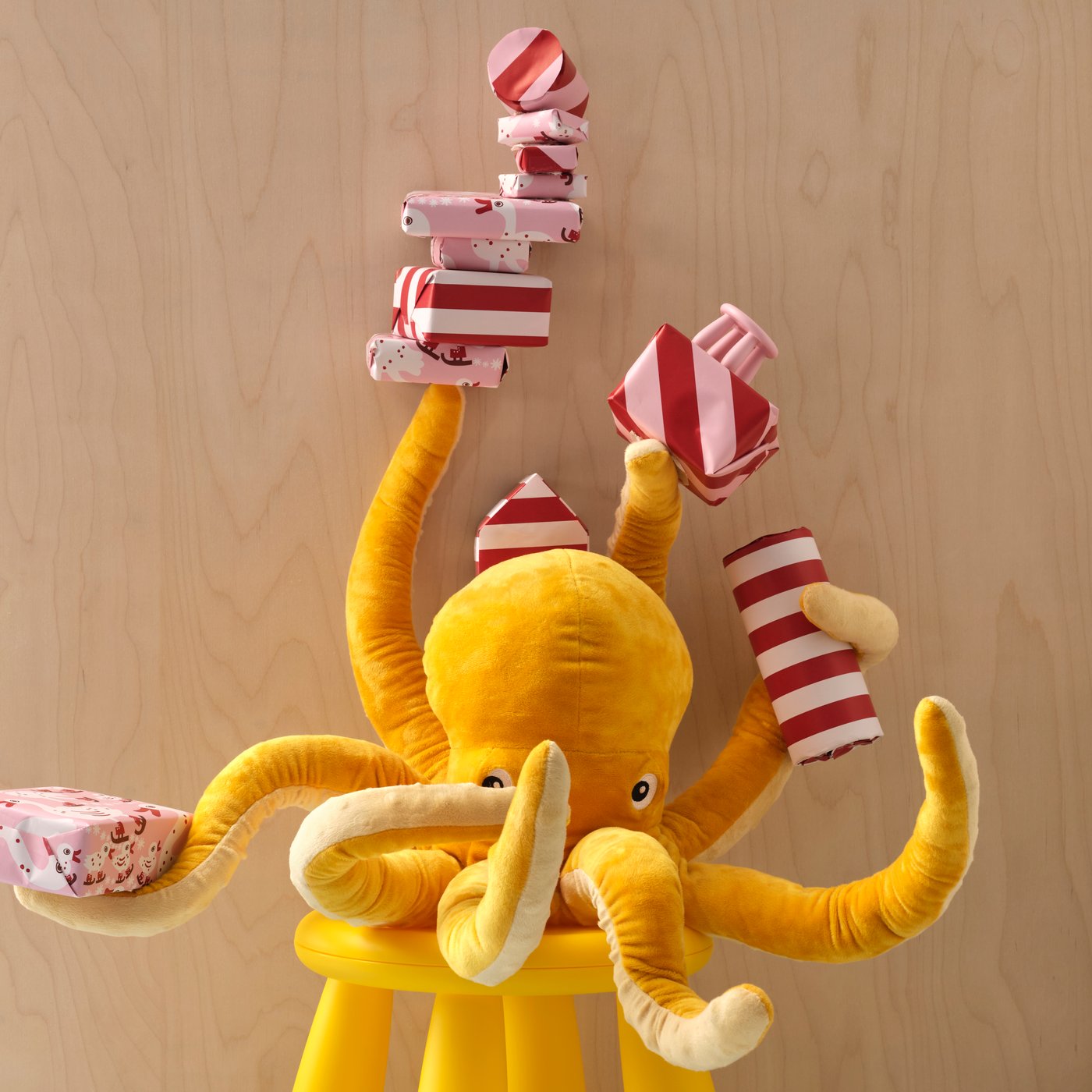 A yellow BLÅVINGAD soft toy octopus is sitting on a MAMMUT stool, holding several wrapped gifts in five of its eight arms.