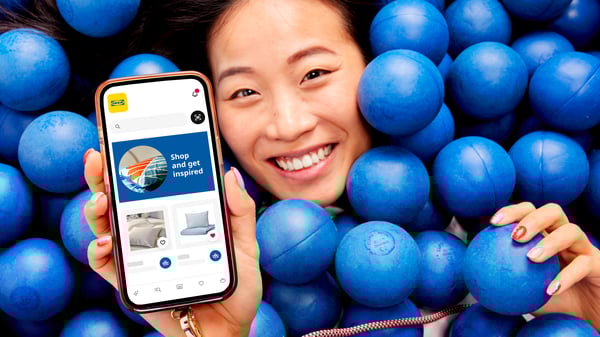 A person lies among blue balls, holding a smartphone that displays the IKEA app.