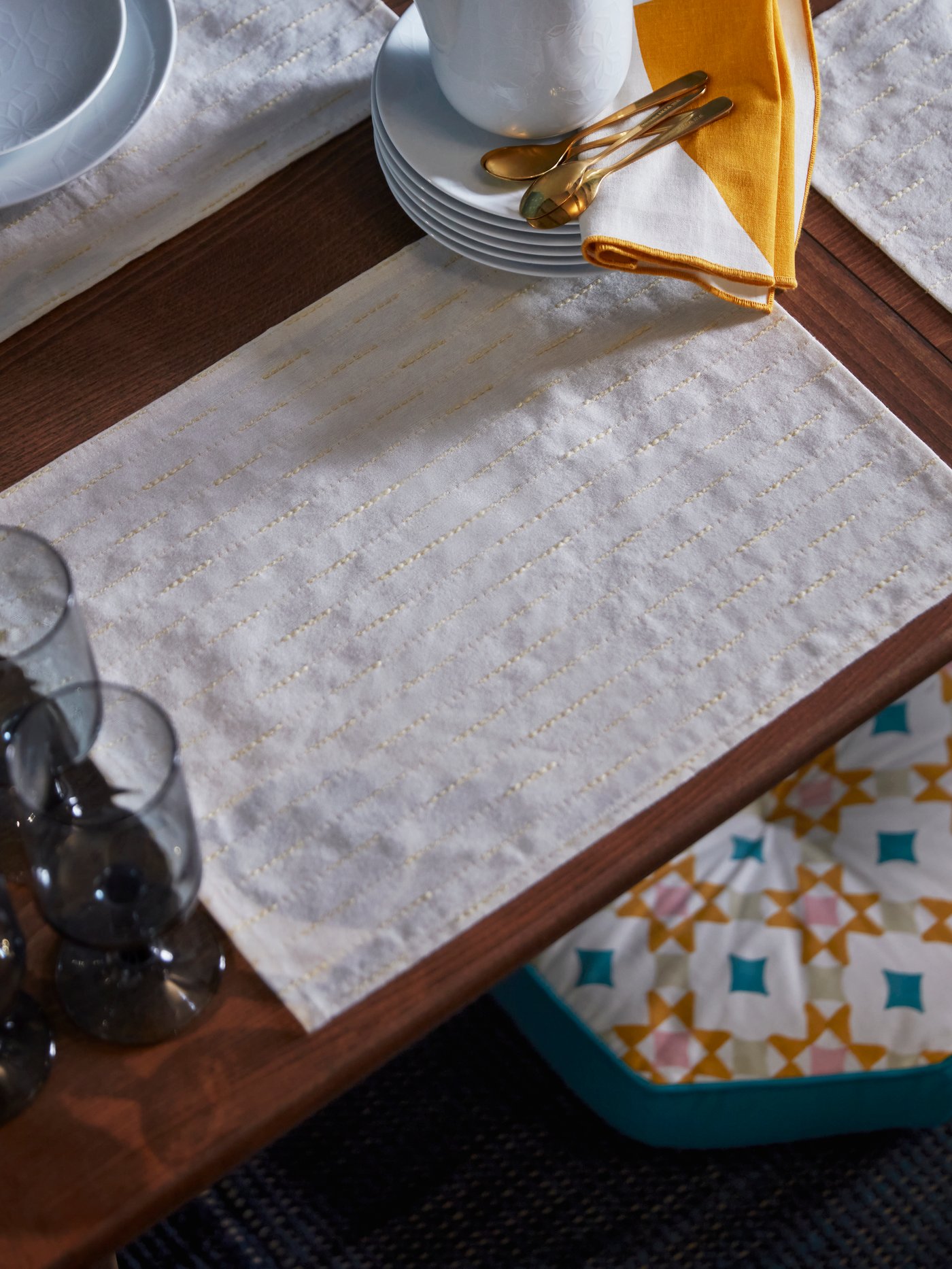 Primer pla d’un parament de taula amb estovalles individuals blanques amb detalls daurats, plats, coberts daurats amb tovalló groc i un coixí de terra amb estampat geomètric de la col·lecció HÖSTAGILLE.