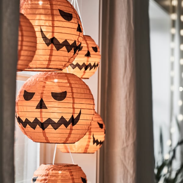 Some KUSTFYR pendant lamps resembling carved pumpkins are hanging in front of a window, next to a curtain.