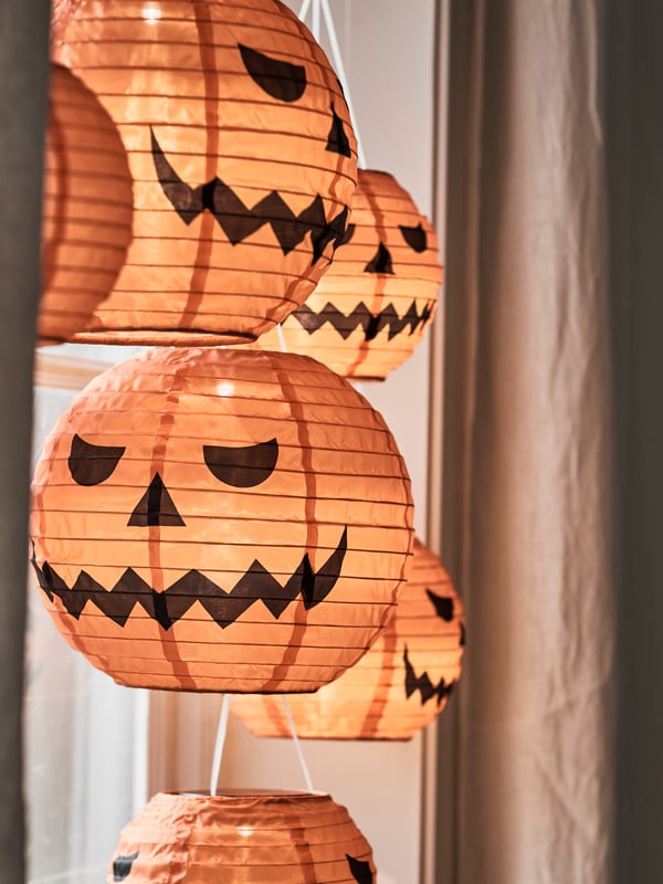 Some KUSTFYR pendant lamps resembling carved pumpkins are hanging in front of a window, next to a curtain.