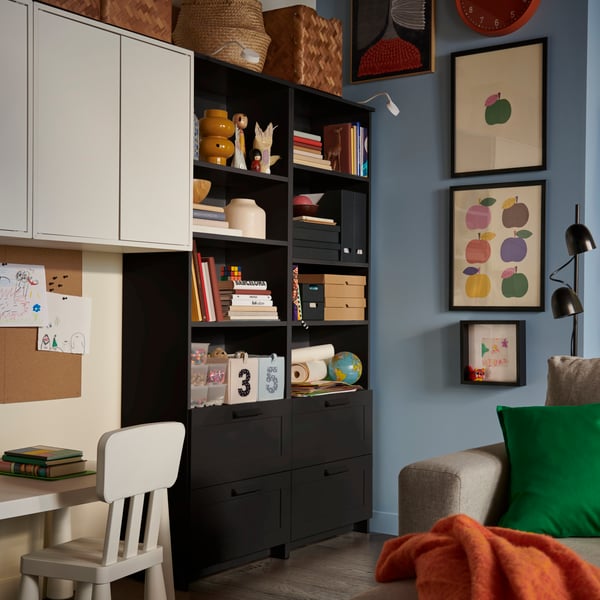 Une bibliothèque BRIMNES noire chargée de livres et de boîtes décoratives, à proximité d’un bureau pour enfant avec une chaise assortie.