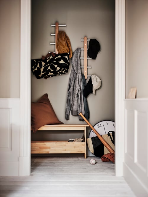 Hallway area featuring two wall-mounted, vertical hook racks with clothes and caps, a bench, a cushion and a bag.