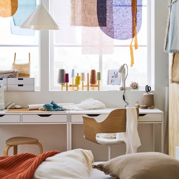 Two white ALEX desks in a sunny bedroom placed side by side create a hobby area with a FJÄLLBERGET conference chair in front.