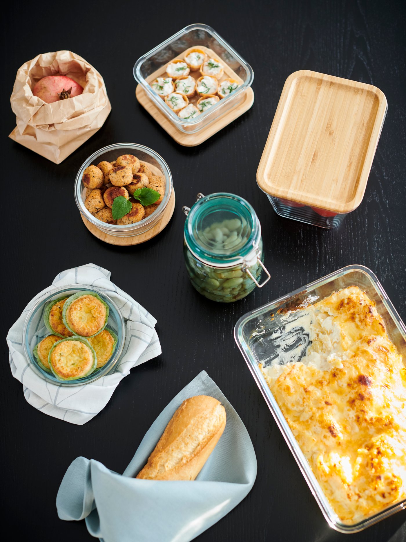 Mesa negra vista desde arriba con recipientes de vidrio para alimentos, algunos con tapas de bambú, y un tarro verde con tapa.