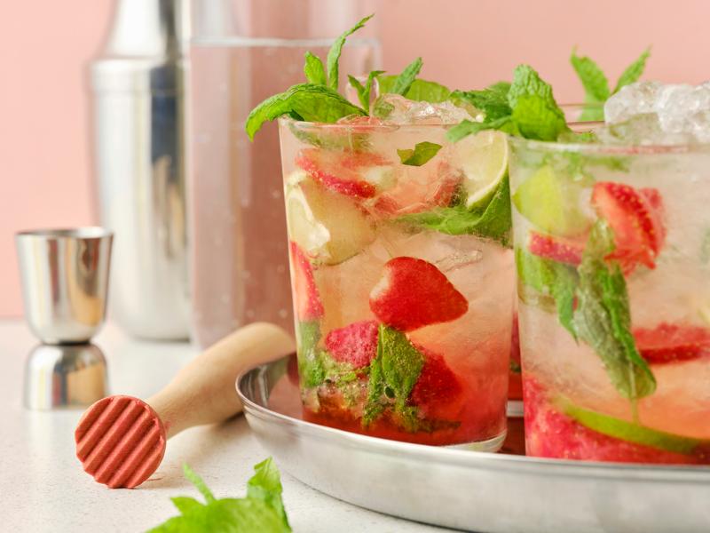 Bicchieri di bevanda gassata al gusto di limone e lime ISKUB, fragole e menta serviti su un vassoio d'argento con utensili da cocktail a lato.