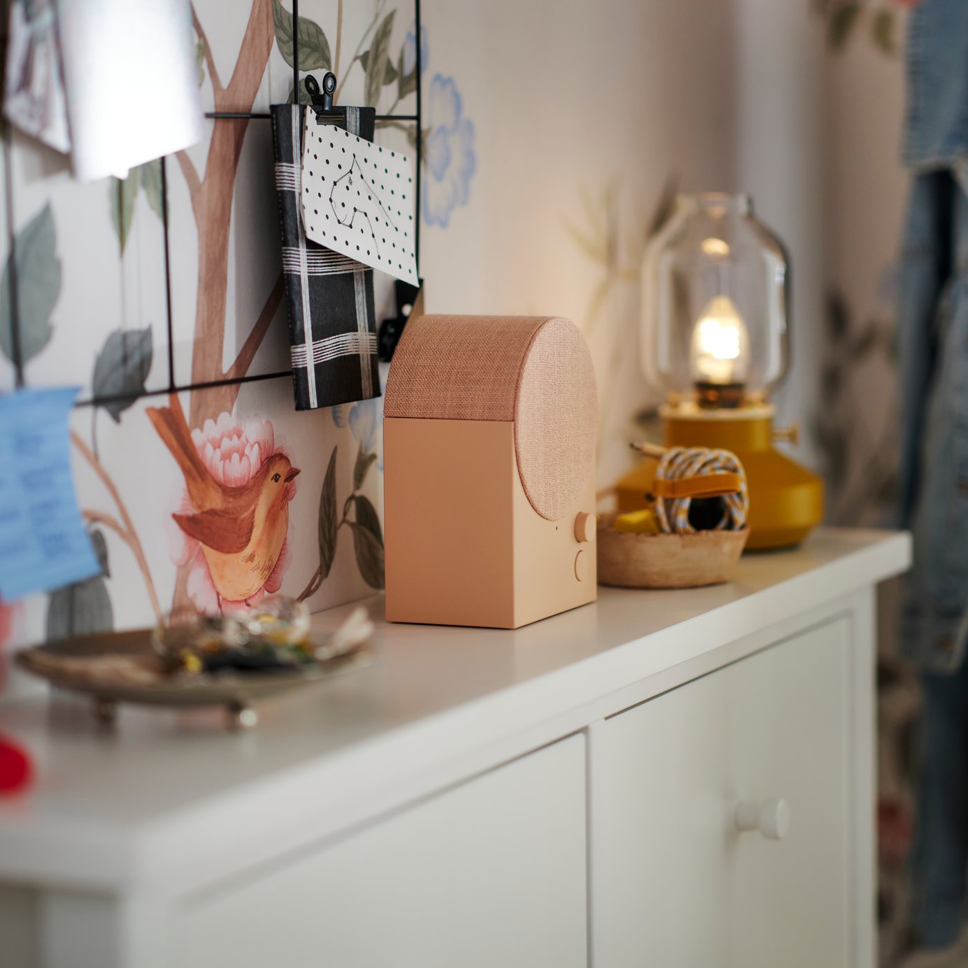 En rosa NATTBAD Bluetooth-högtalare ovanpå ett vitt GULLABERG skoskåp mellan en bordslampa och ett fat med nycklar i.