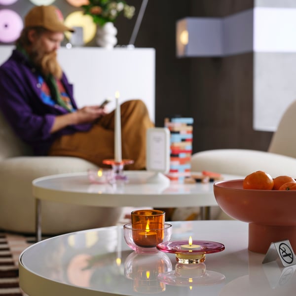 A white table has a glass, pink/brown TALLTITA tealight holder set with a candle by other decor in a modern hotel lobby.