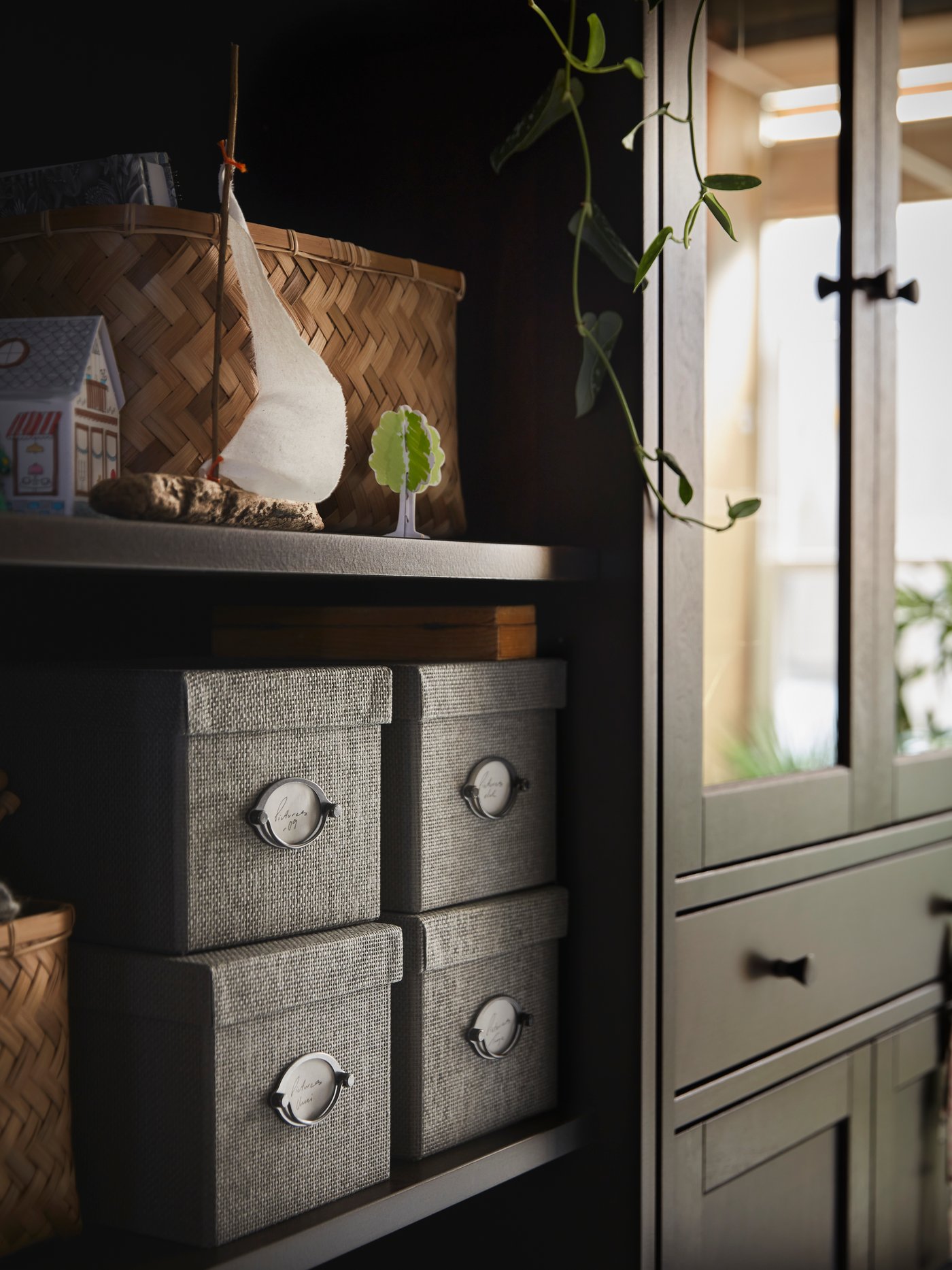 Four KVARNVIK boxes stand on a shelf of an IDANÄS bookcase which stands beside a high cabinet with glass-doors and 1 drawer.