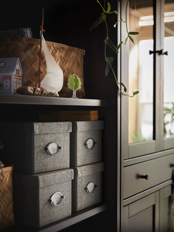 Four KVARNVIK boxes stand on a shelf of an IDANÄS bookcase which stands beside a high cabinet with glass-doors and 1 drawer.