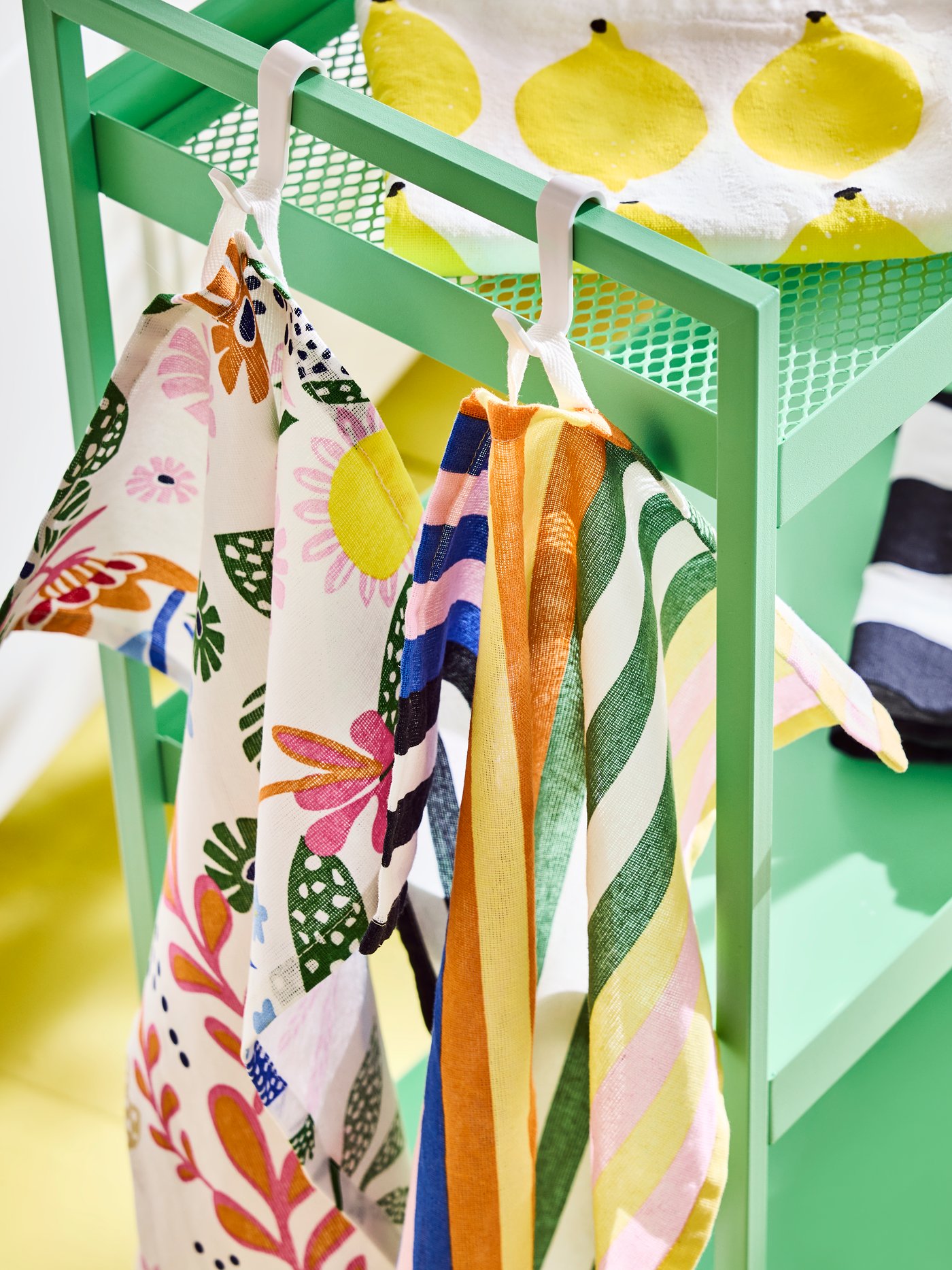 Set of four colourful RINNIG tea towels with lemon, flower and striped motifs hanging from a light green NISSAFORS trolley.