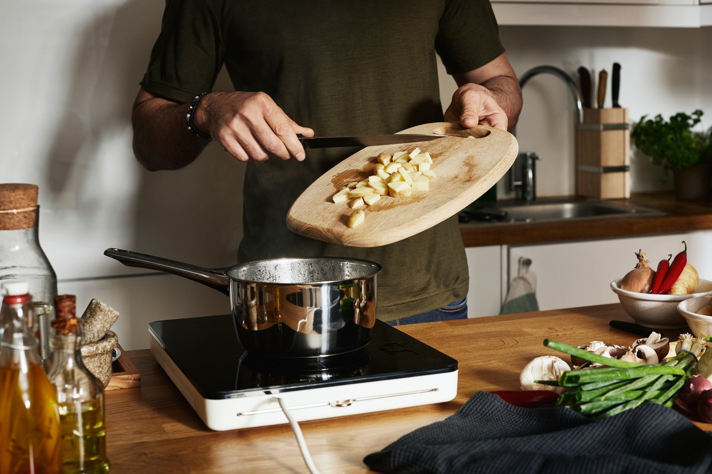 A person is dropping chopped potatoes from a chopping board into a chrome casserole on a TILLREDA portable induction hob.