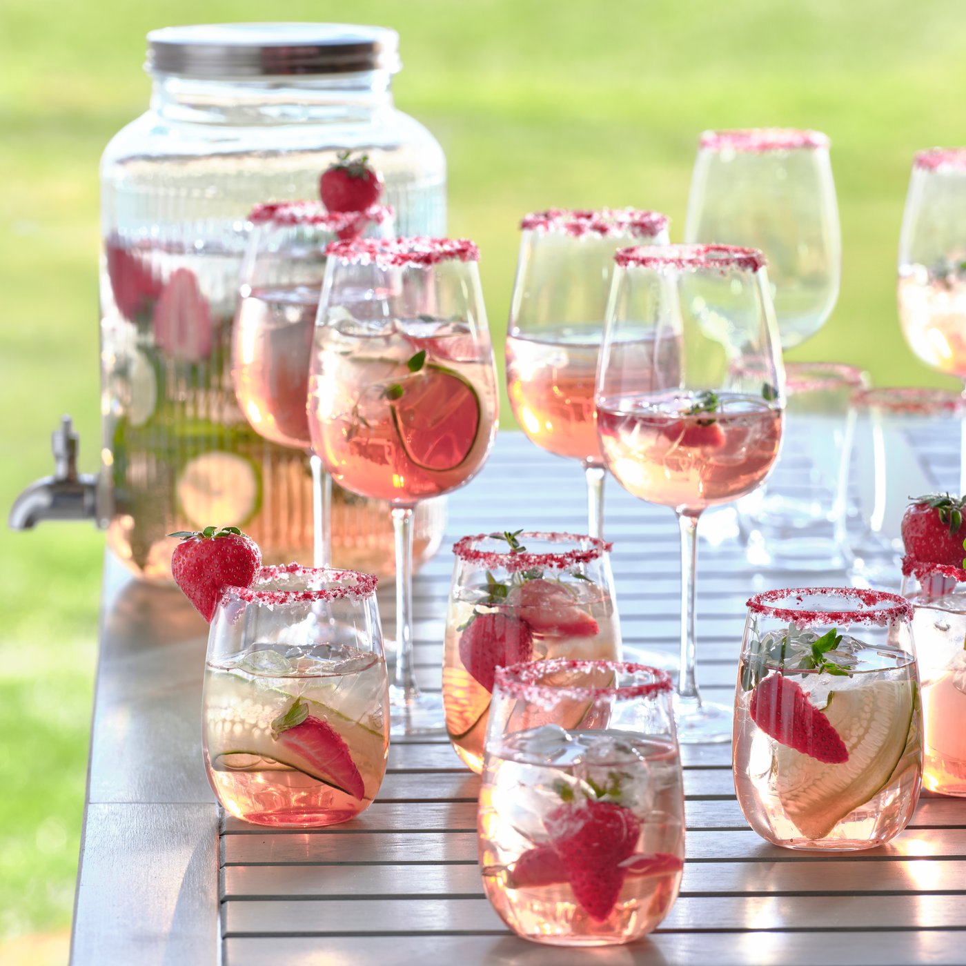 Glas med rosa dryck, jordgubbar och is står på ett träbord utomhus, med en stor dryckesbehållare i bakgrunden