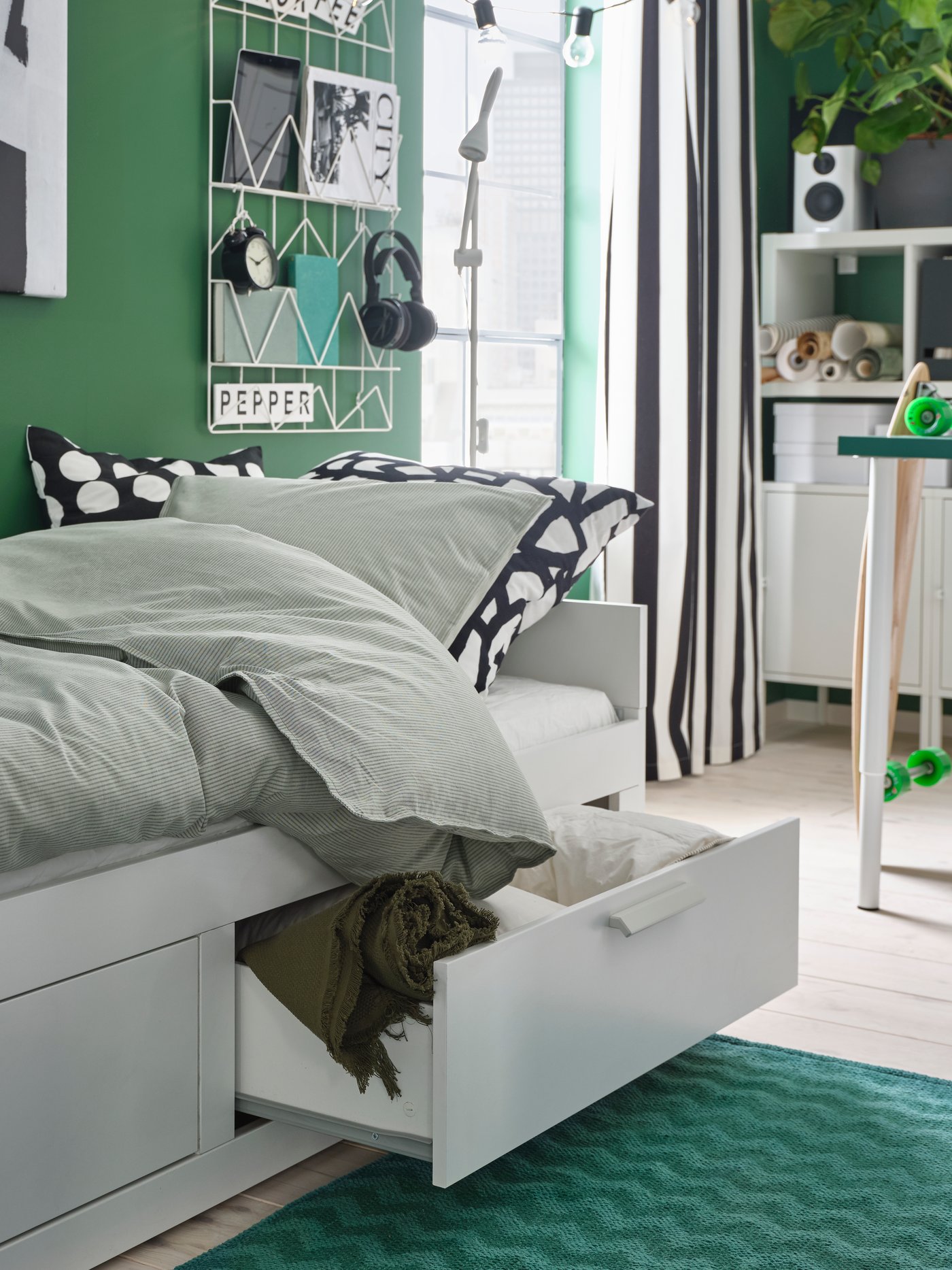 Dormitorio con paredes verdes, alfombra verde, estructura de cama en blanco con dos cajones, módulo de almacenaje blanco y lámpara de pie.