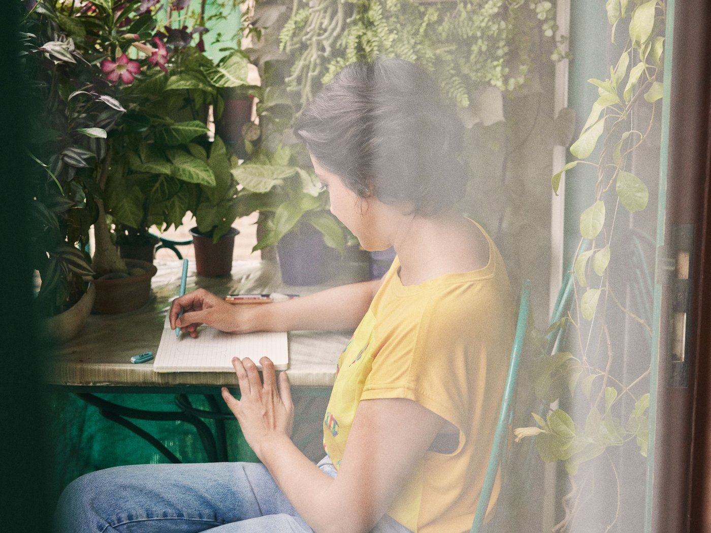 A woman in a yellow shirt sits at a table in her garden and writes in her book.