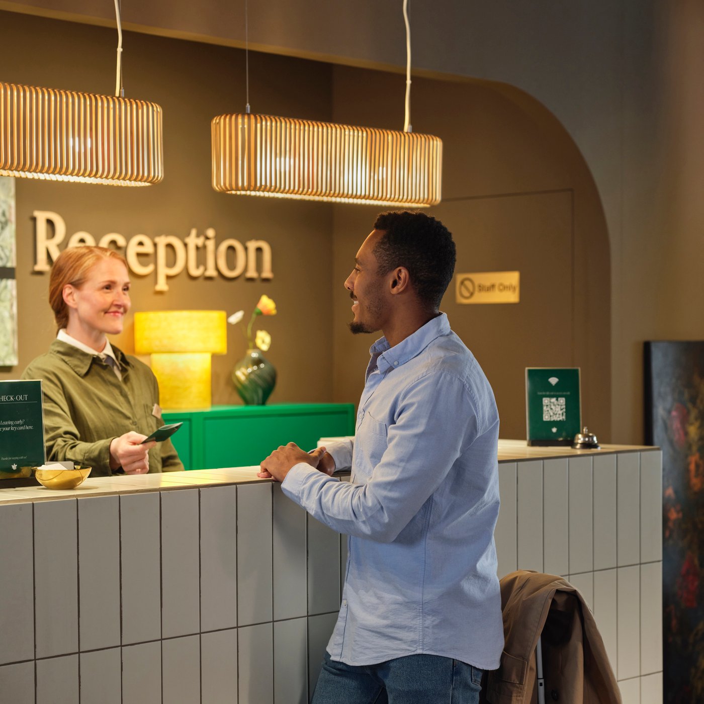 Hotellreception; personalen ger gästen ett visitkort under varma taklampor, skylten ”Reception” syns bakom disken.