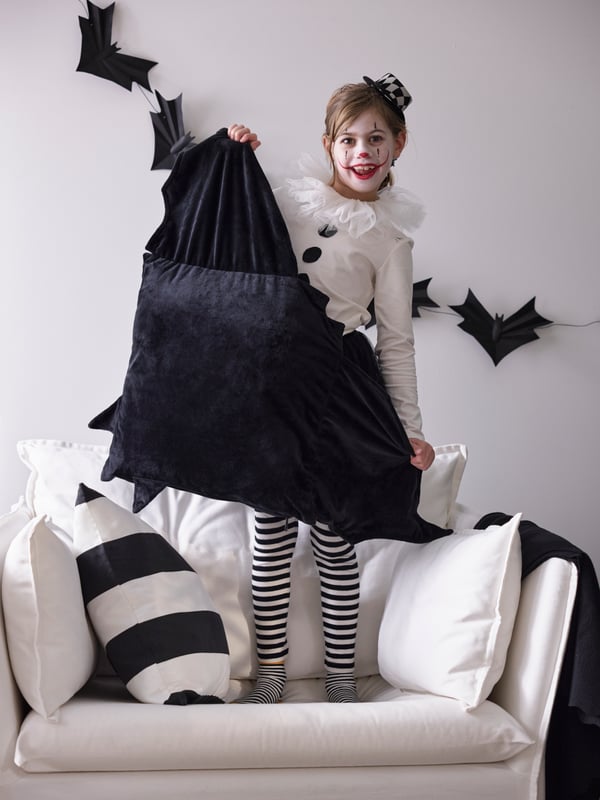 A child dressed in a clown costume stands on an armchair while holding a HÖSTAGILLE bat-shaped cushion.