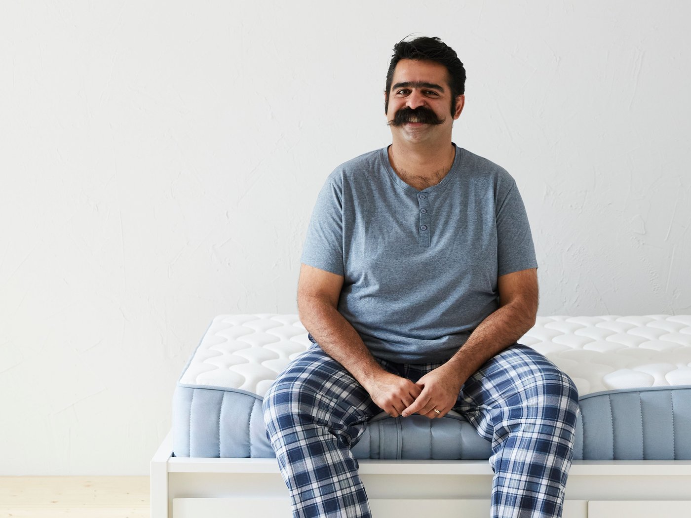 شخص له شارب يرتدي قميصاً باللون الأزرق المائل إلى الرمادي وبنطلون بيجامة بنقش المربعات يجلس على حافة مرتبة.