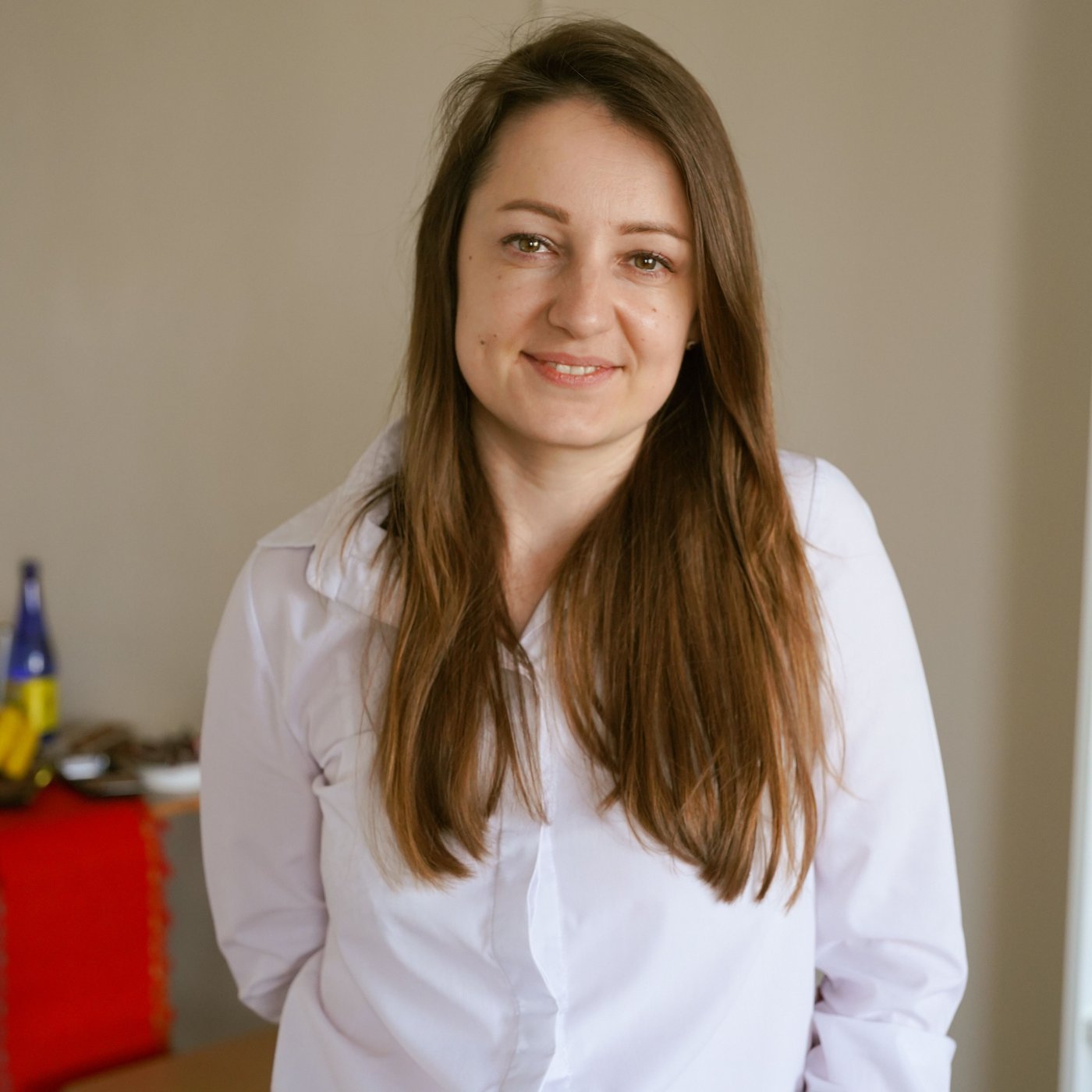 Eine junge Person steht in einem Raum und schaut in die Kamera. Sie trägt ein weisses Hemd und hat langes braunes Haar.