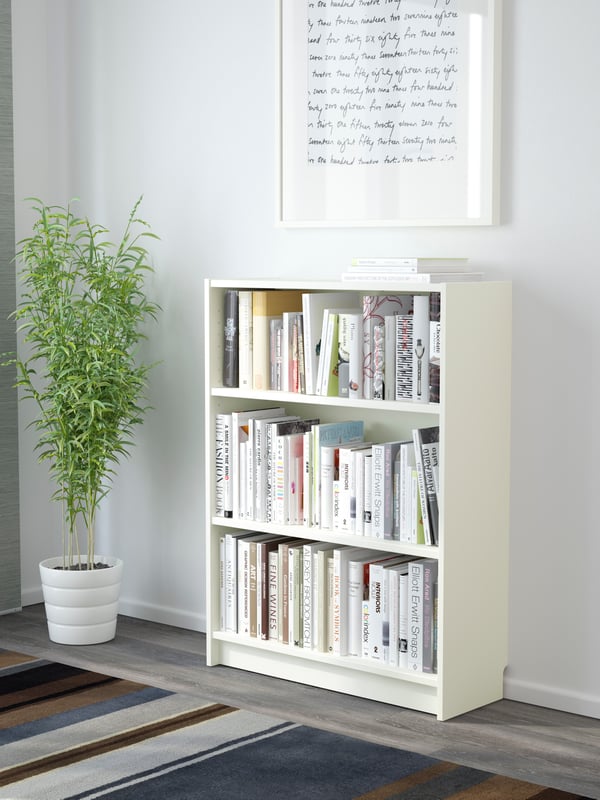 Ein weißes BILLY Bücherregal  80 breit, 28 tief und 106 cm hoch, steht vor einer weißen Wand. Daneben steht eine Zimmerpflanze. 
