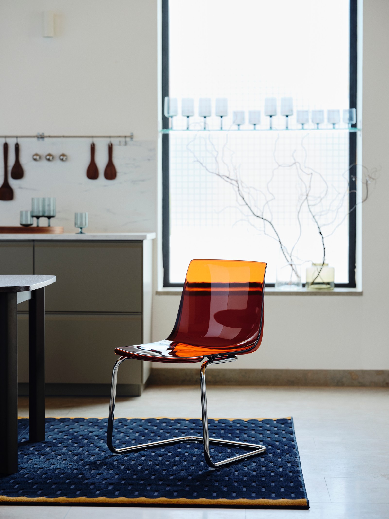 A TOBIAS chair in brown/red/chrome-plated steel sits in front of a window on a navy textured rug in a bright kitchen.
