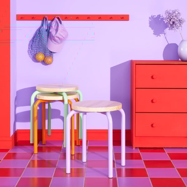Es ist ein Zimmer in Lila und Rot mit einem Schachbrettboden in den gleichen Farben zu sehen. Im Raum befinden sich IKEA Hocker, eine Kommode und Kleiderbügel.
