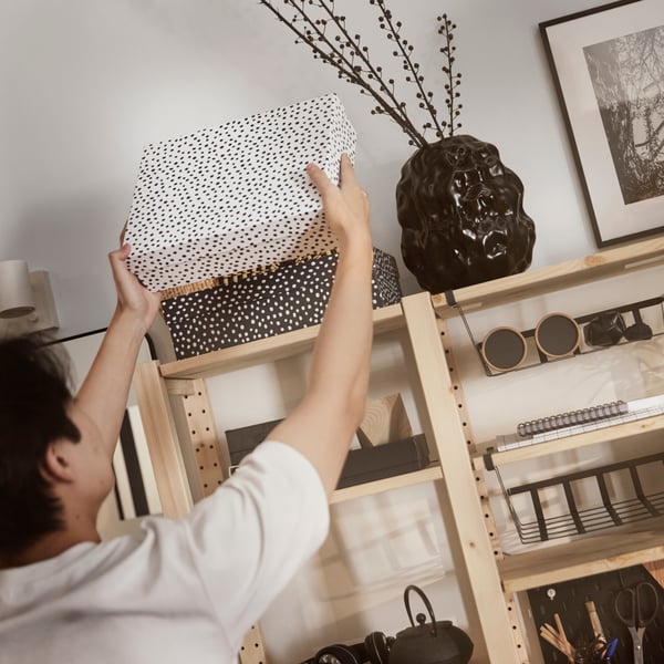 A person placing a RÄTTAREN storage box in white with black dots in an IVAR shelving unit in pine with black decoration.