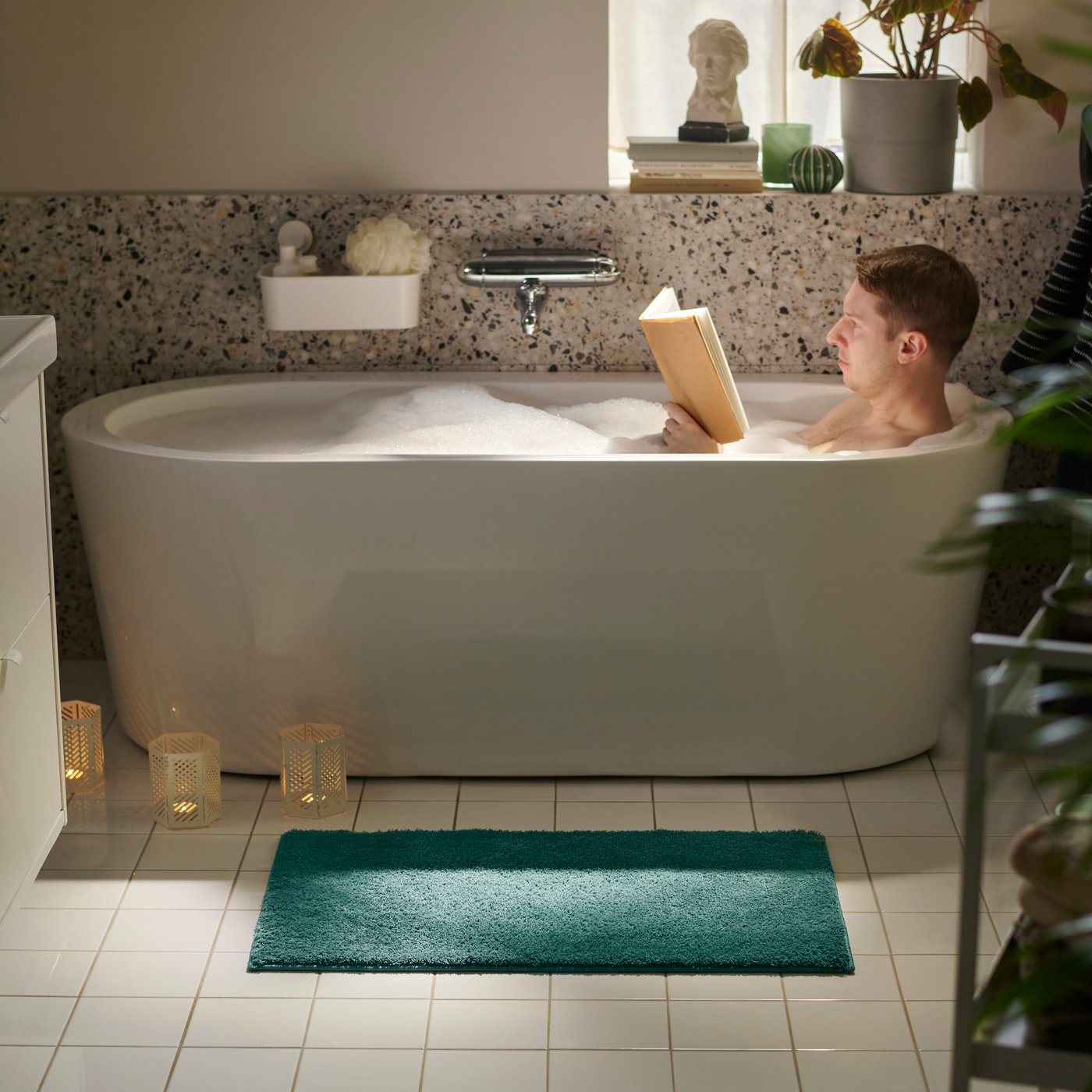 Et baderom med en BACKMÅRA badematte på gulvet ved siden av badekaret, der en person slapper av i badet.