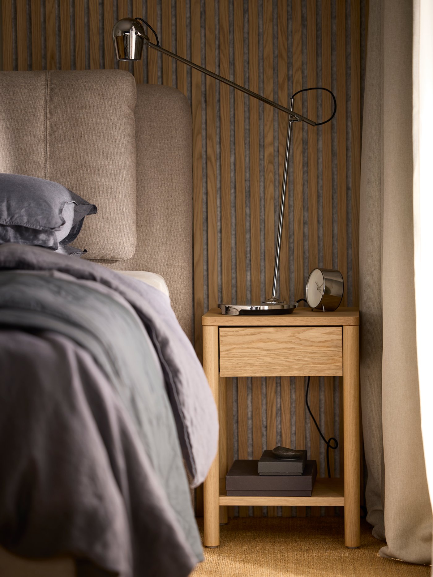 An oak veneer TONSTAD bedside table with a chrome-plated MODERMOLN work lamp on top, by a LILLFJÄLLET divan bed with storage.