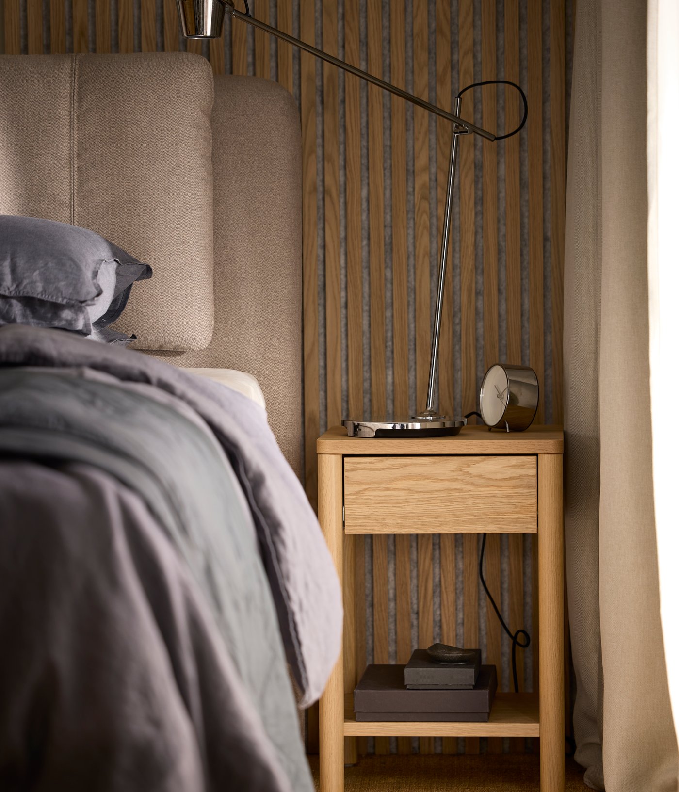 An oak veneer TONSTAD bedside table with a chrome-plated MODERMOLN work lamp on top, by a LILLFJÄLLET divan bed with storage.