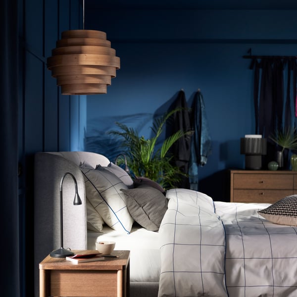 Against a dark blue wall, a white stained oak veneer DYKARKLOCKA pendant lampshade hangs above a TUFJORD upholstered bed.