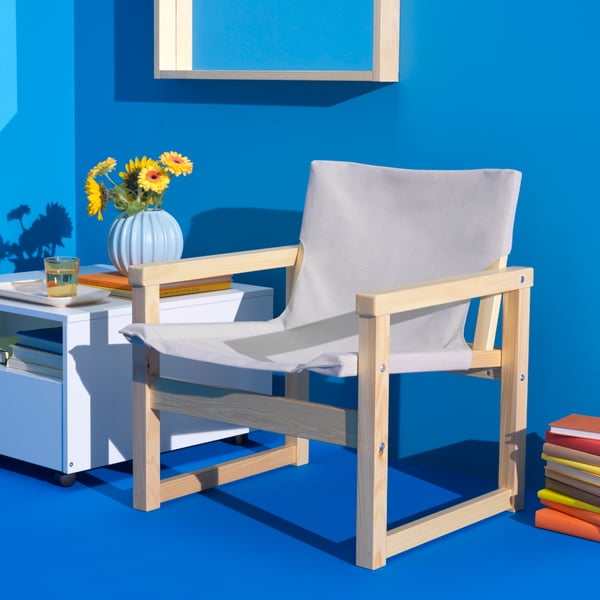 A SJÄLSÖ armchair made of solid pine frame and a strong natural-coloured fabric in the seat and back sits in a blue-painted room. A bunch of sunflowers are in a blue SKOGSTUNDRA vase, sitting atop of a white GARNANÄS coffee table.