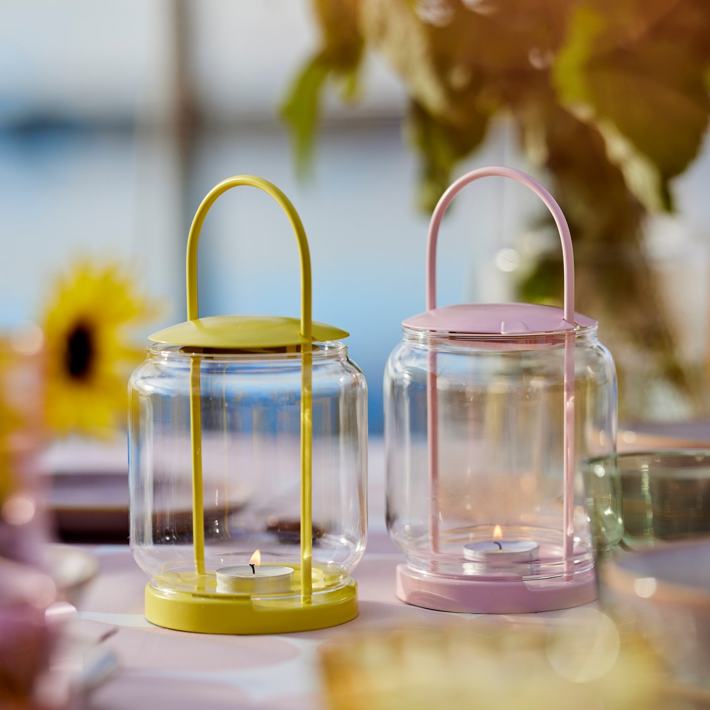 A dining table outdoors decorated with two minimalistic TOFSAND lanterns in pink and yellow with tealights burning inside.