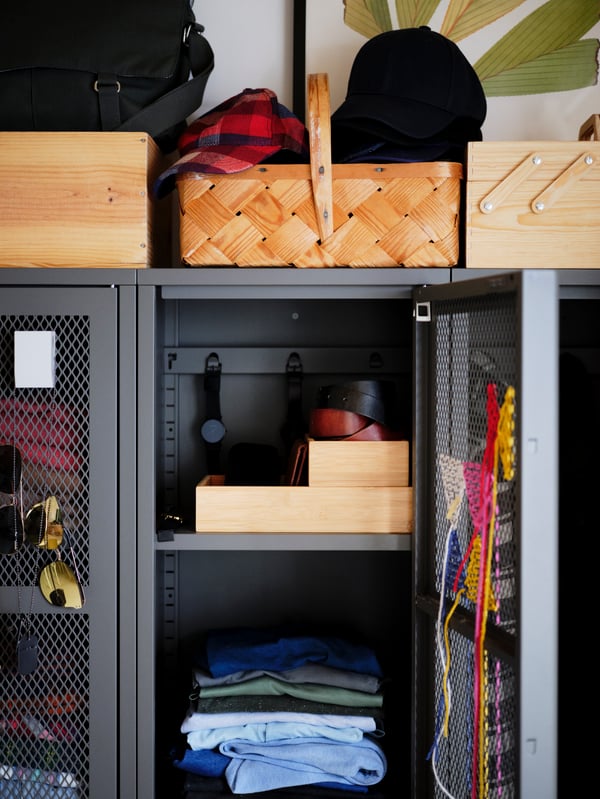 A section of a grey IVAR cabinet holding clothes and accessories. There's embroidered decorations on the door's metal mesh.